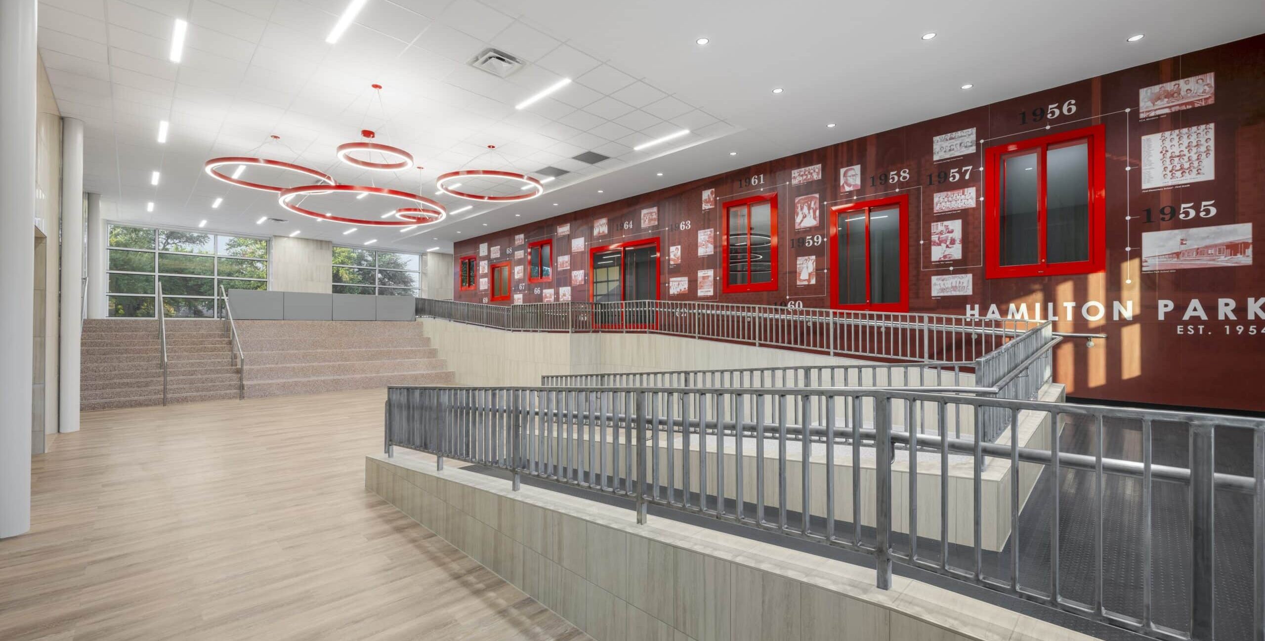 A spacious interior area with light wood flooring, a ramp with metal railings, and tiered seating steps. A large wall displays a historical timeline with photos and dates, along with the text “Hamilton Park Est. 1954.” Red-framed windows and circular red ceiling lights add contrast, and natural light enters through tall windows on the left.