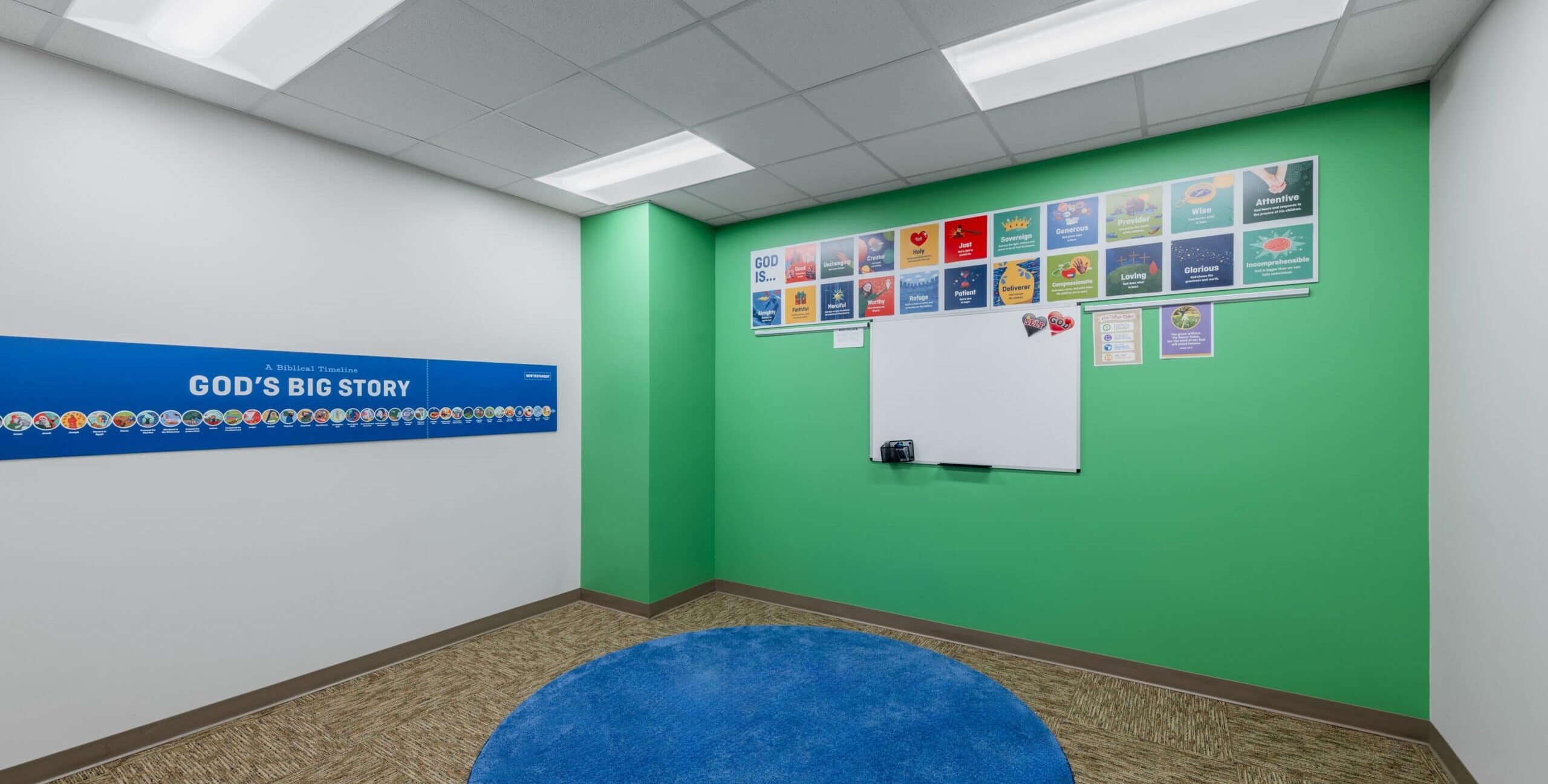 A small classroom with bright green and white walls, a large blue circular rug, and a long blue “God’s Big Story” timeline banner on the left wall. A whiteboard is mounted on the green wall beneath colorful “God Is…” attribute posters.