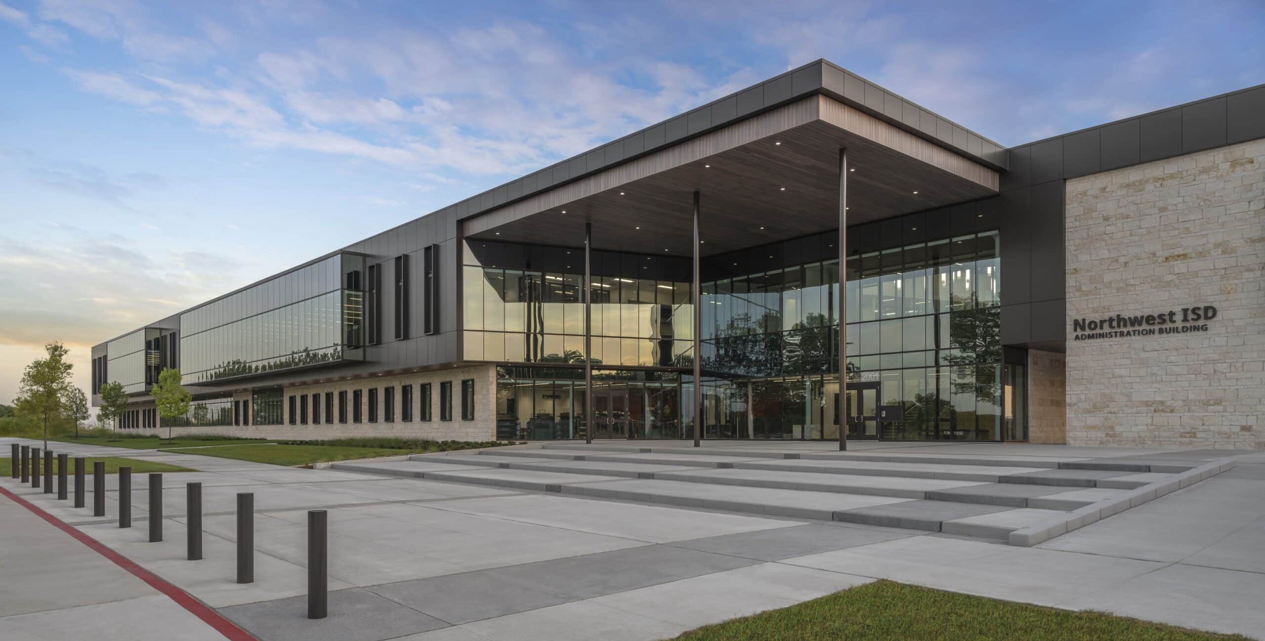 A modern two‑story administration building with large glass windows, stone and metal exterior panels, and a wide entrance with steps leading to glass doors under an extended canopy. The surrounding area includes paved walkways, bollards, grass, and young trees.