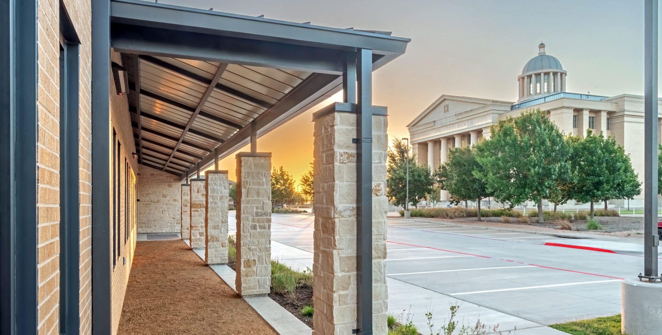 A sunrise view of a covered walkway supported by stone columns and a metal roof beside a brick wall. In the background stands a neoclassical building with a dome and columns, illuminated by warm evening light.