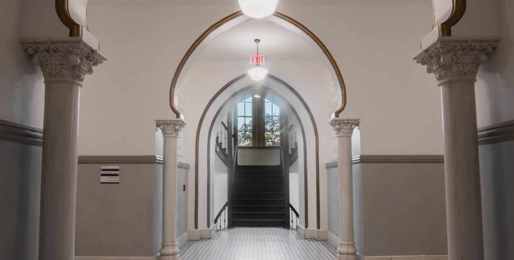 A long interior hallway with Gothic-style pointed arches trimmed in gold and supported by marble columns with ornate capitals. The floor has a repeating hexagonal tile pattern, and hanging light fixtures illuminate the space. At the far end, an exit sign hangs near a staircase leading upward, with a large window providing natural light.