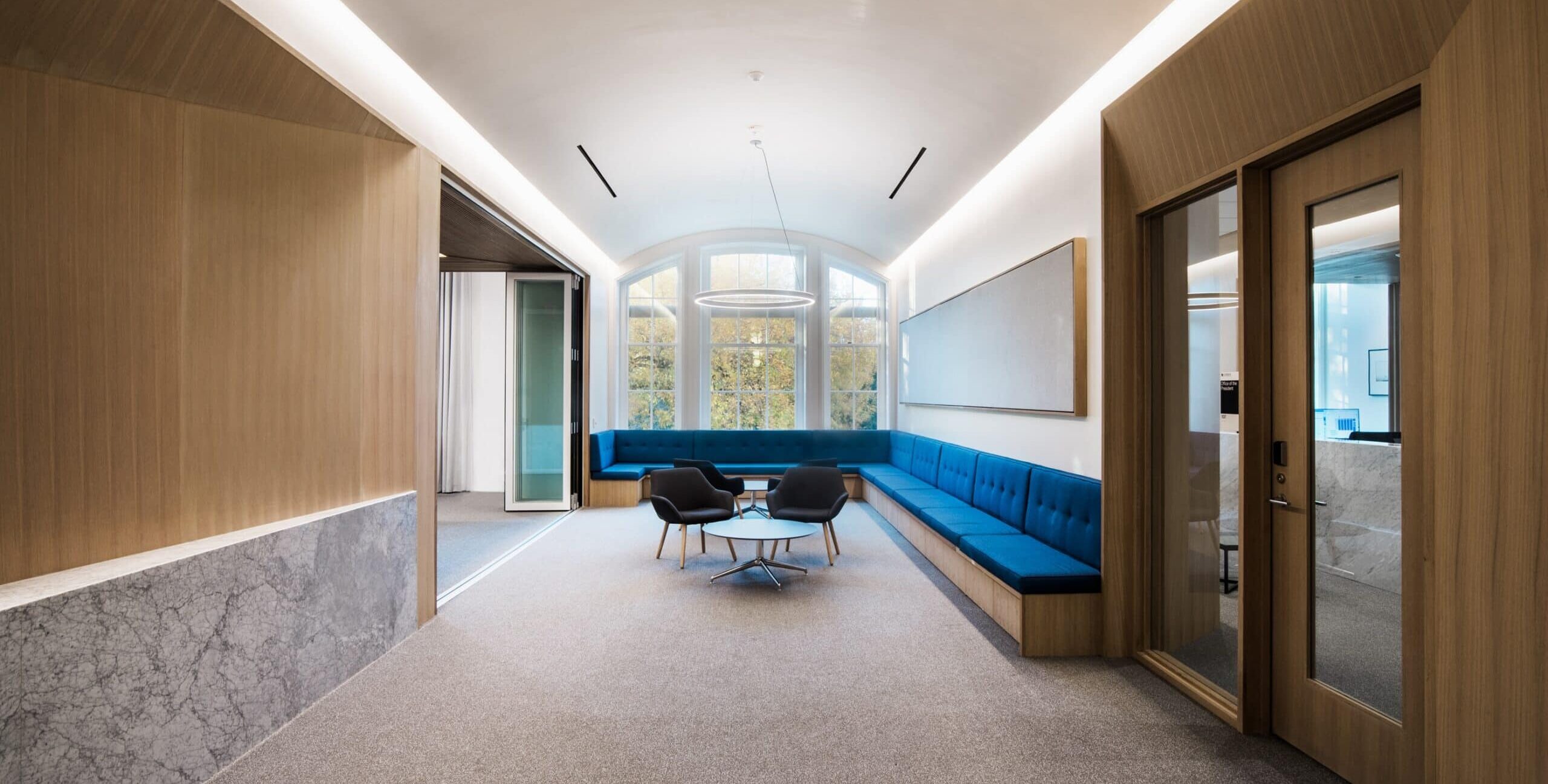 A modern office lounge with an L‑shaped blue upholstered bench along two walls and a small round table with three dark gray chairs in the center. Large arched windows at the far end let in natural light and show trees outside. The space features white walls, wood paneling and marble accents on the left, and glass doors to adjacent rooms on the right. Recessed linear lighting brightens the room.