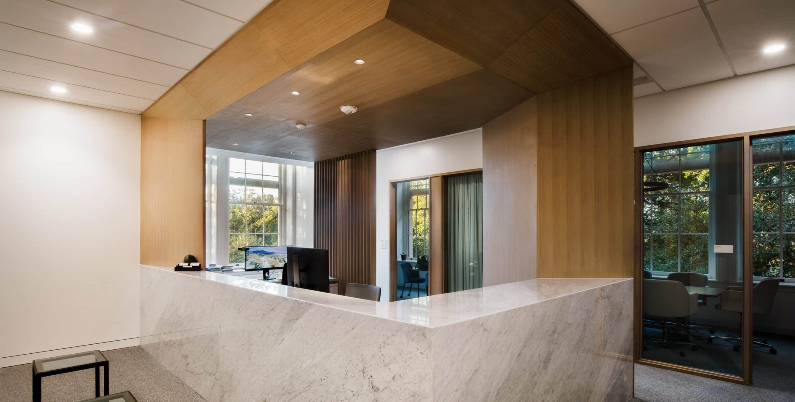 A modern office reception area with a large L‑shaped marble desk and a computer setup behind it. Wood paneling covers the ceiling and part of the walls, contrasting with white walls and marble surfaces. To the right, glass windows and doors reveal a meeting room with chairs and a view of greenery outside. In the foreground, two small glass‑top tables sit on a gray carpet. The space is bright with recessed lighting and natural light from the windows.
