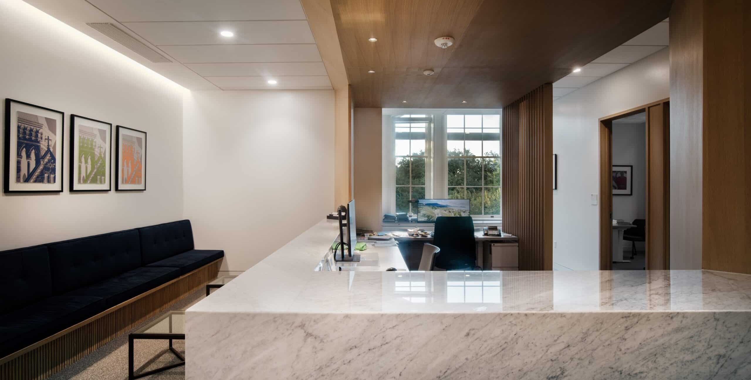 A modern office reception area with a large marble counter in the foreground. Behind the counter is a workspace with a computer, office chair, and desk items. To the left, a long dark‑blue upholstered bench sits beside a minimalist glass coffee table. Above the bench hang three framed artworks in blue, green, and orange. A large window in the back lets in natural light and shows greenery outside. The ceiling features recessed lighting with white and wood panel sections.
