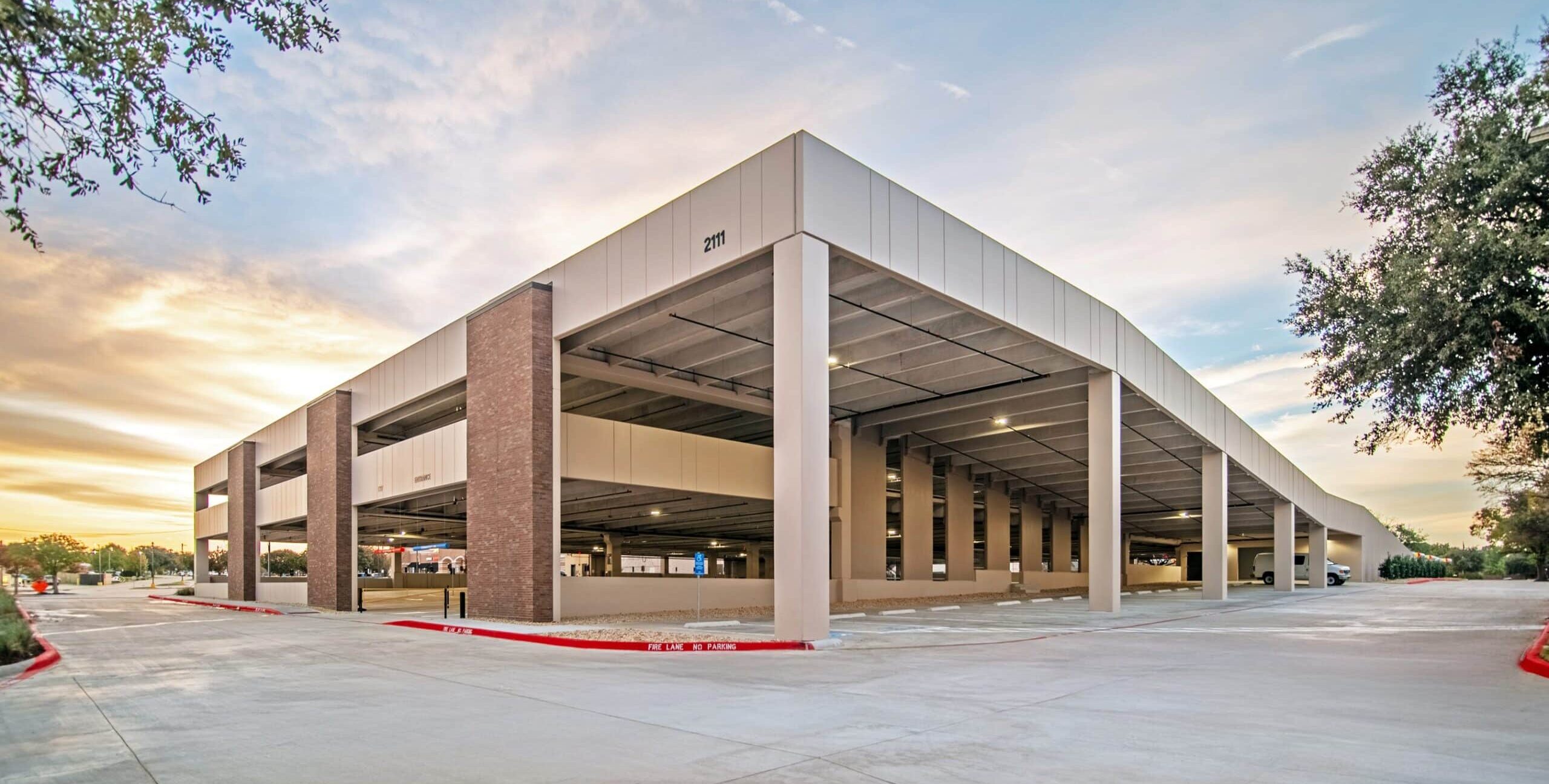 A modern multi‑level parking garage featuring brick and metal panels with large open sections. The number “2111” appears near the roofline. A wide concrete driveway surrounds the building, and a sunset sky with soft clouds glows in the background.