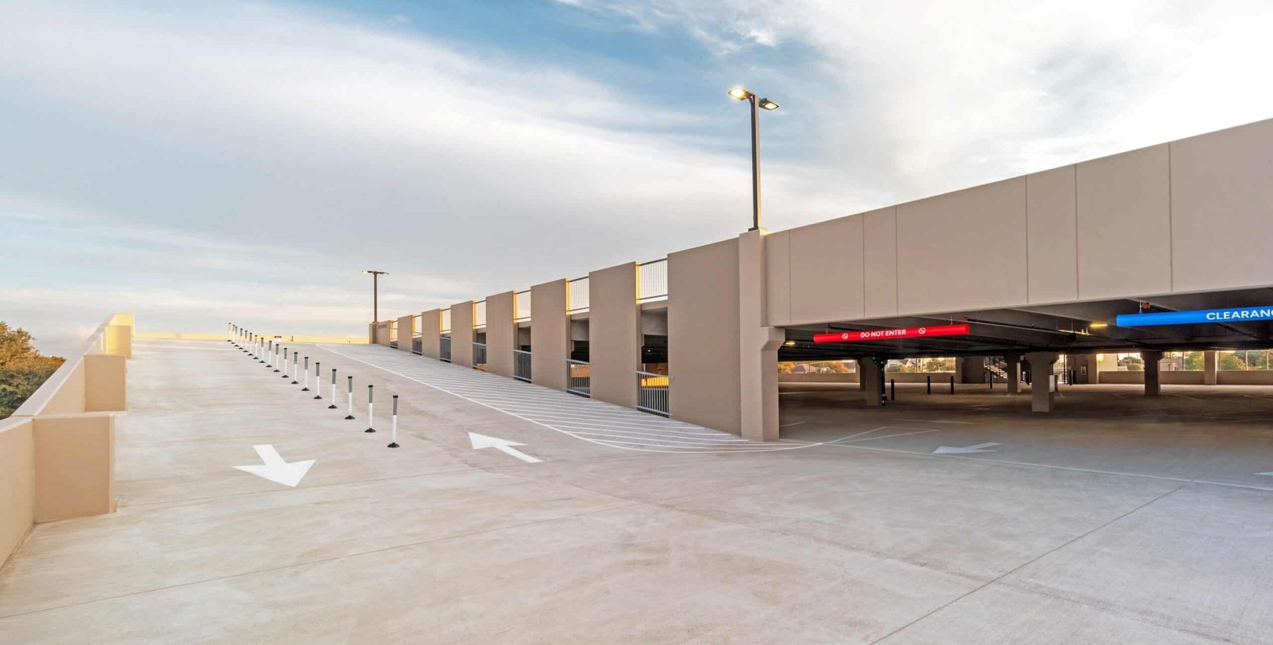 A concrete ramp leading to an upper level of a parking garage. White arrows indicate vehicle direction, and short white posts divide lanes. Overhead signs read “DO NOT ENTER” and “CLEARANCE.” The beige structure is lit by daylight under a partly cloudy sky.