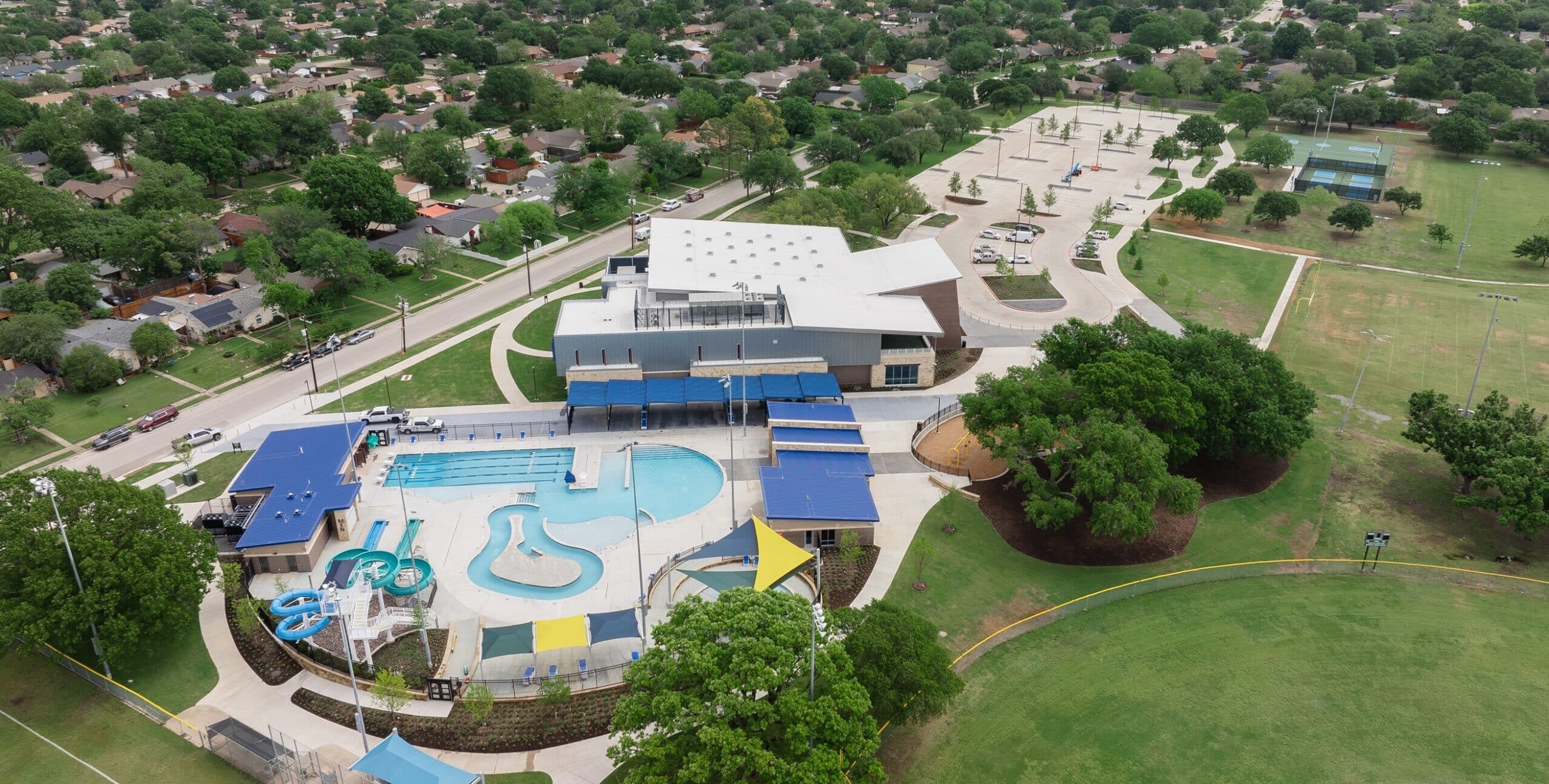 An aerial view of a community recreation center with a large outdoor pool, lazy river, water slides, shaded seating areas, nearby sports fields, pickleball courts, and surrounding residential neighborhoods.