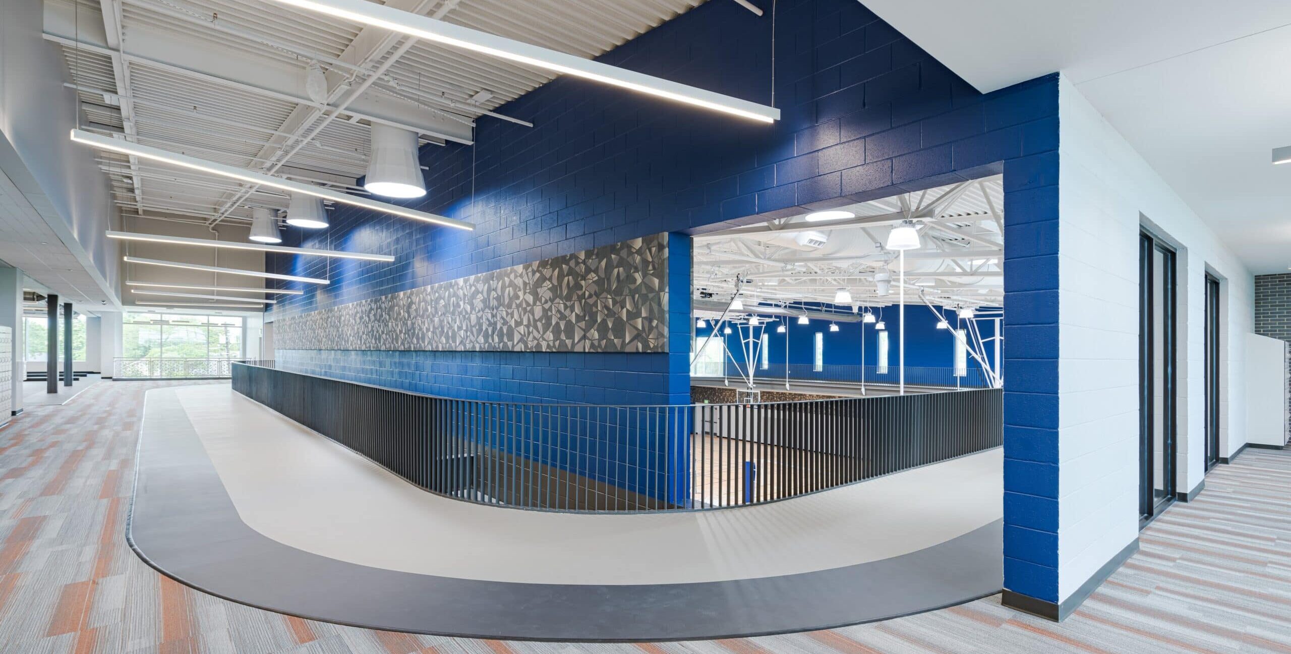 A modern indoor fitness facility with an elevated curved walking track, blue and white walls, patterned accent panels, exposed beams, linear lighting, and large windows providing natural light.
