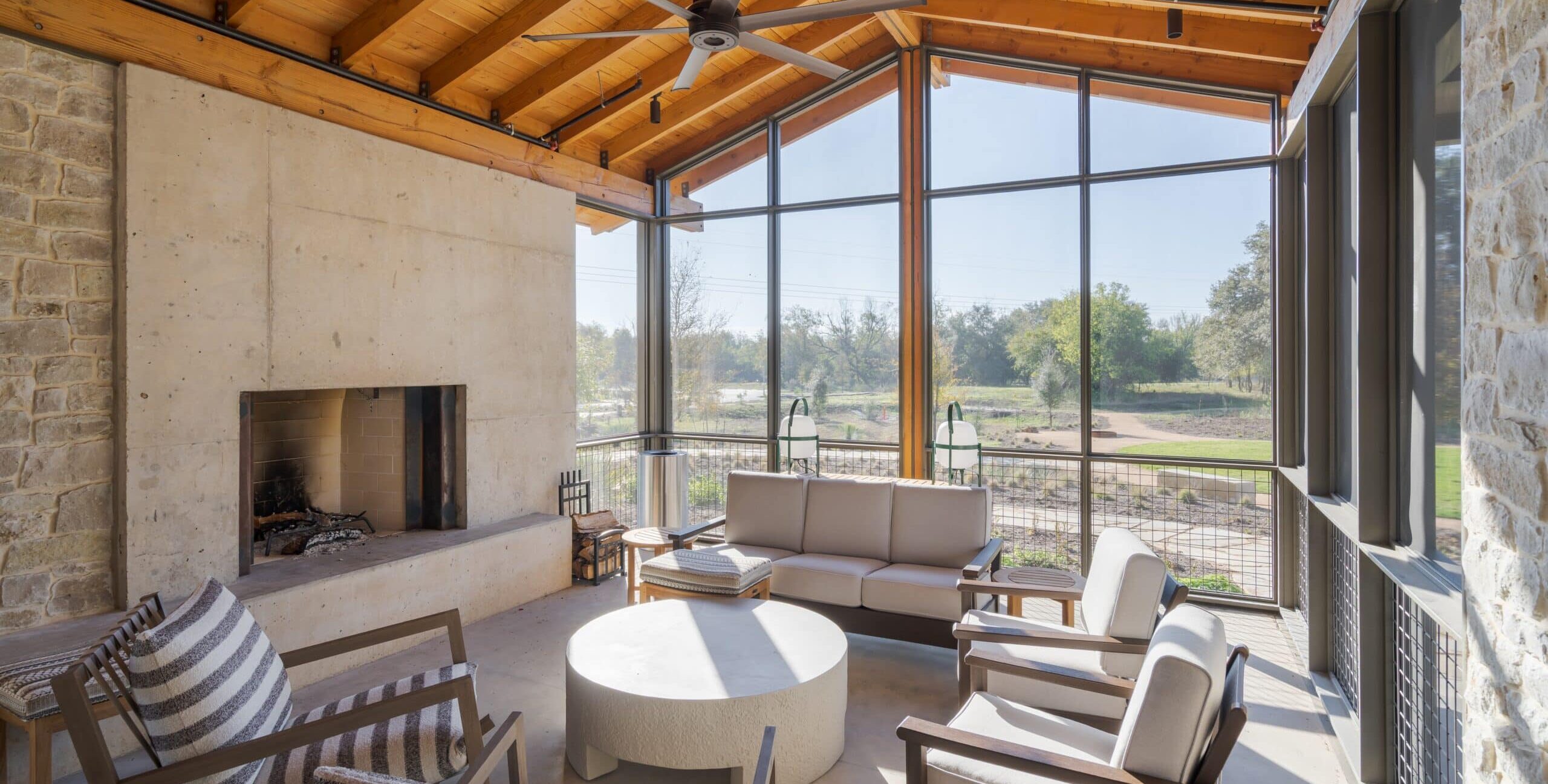 A bright, screened-in porch featuring a large concrete fireplace and floor-to-ceiling windows. The room is furnished with comfortable outdoor armchairs and a circular coffee table under a large industrial ceiling fan.