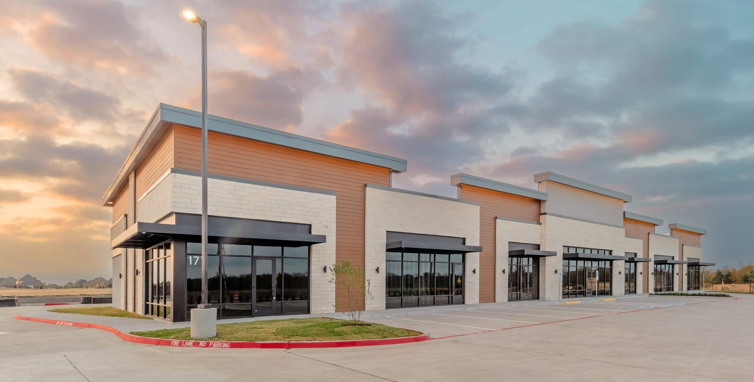 A newly built commercial strip center with large glass storefronts, light stone walls, wood paneling near the roofline, and black metal awnings. A wide, freshly paved parking lot sits in front beneath a sunset sky.