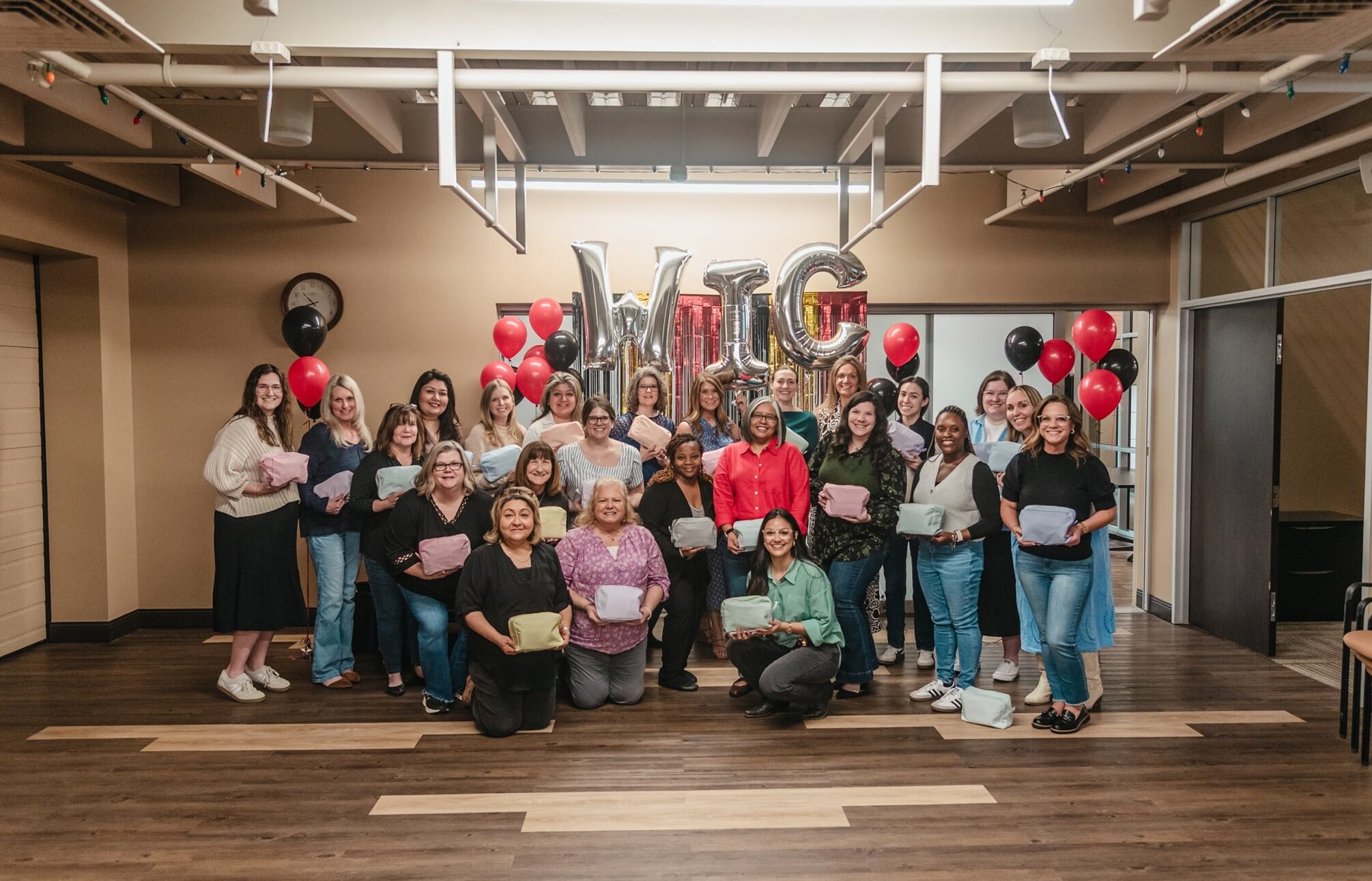 A group of people pose together in a room with wooden floors and beige walls, each holding a small pastel-colored pouch. Behind them, large metallic balloon letters spell “WIC,” surrounded by red and black balloons. A clock hangs on the wall above them, and exposed ceiling beams and lights are visible. The group is smiling and arranged in standing and kneeling rows, suggesting a celebratory or community event.